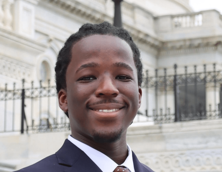 a young Black man standing in front of the United States Capitol while wearing a suit and tie