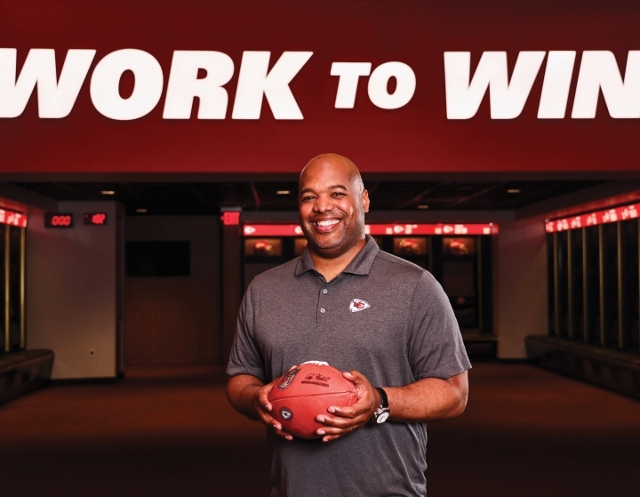 a young Black man holding a football in front of a wall that reads "work to win"