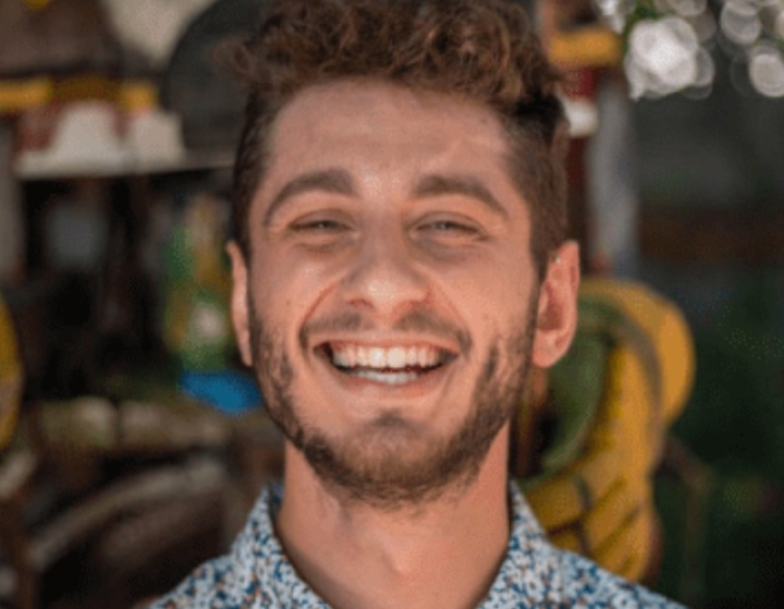 A close-up portrait of a smiling man with curly brown hair and a short beard, wearing a blue and white floral patterned button-down shirt.