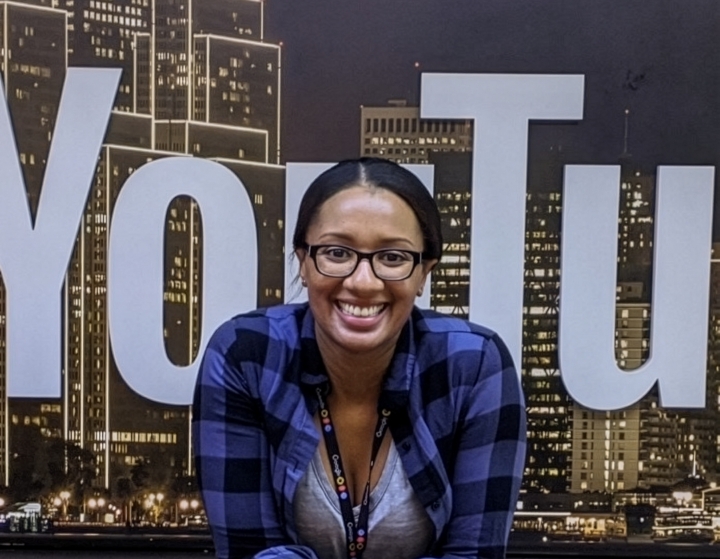 A smiling woman with elbows on a newsroom desk and the YouTube logo behind her
