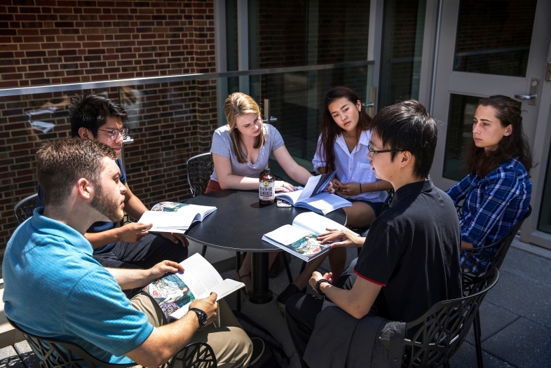 Group of students sits around a round table holding books and talking while on a patio