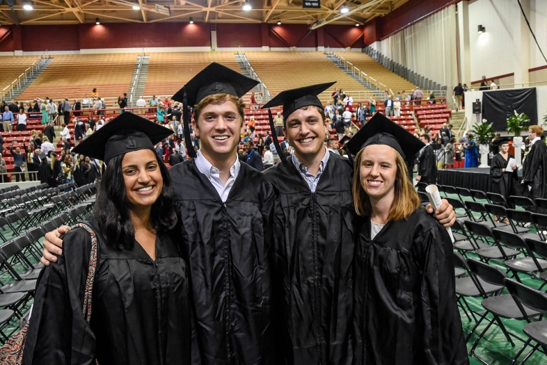 Students smile in caps and gowns at indoor Commencement
