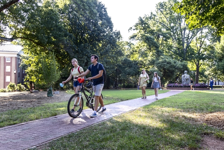 Two students walking, one holding a bike and talking on campus