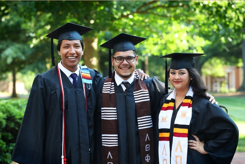 three students standing and smiling in graduation robes