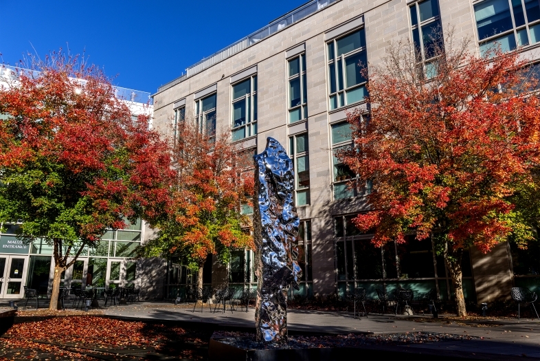 a silver piece of art stands in front of a modern academic building as red trees surround