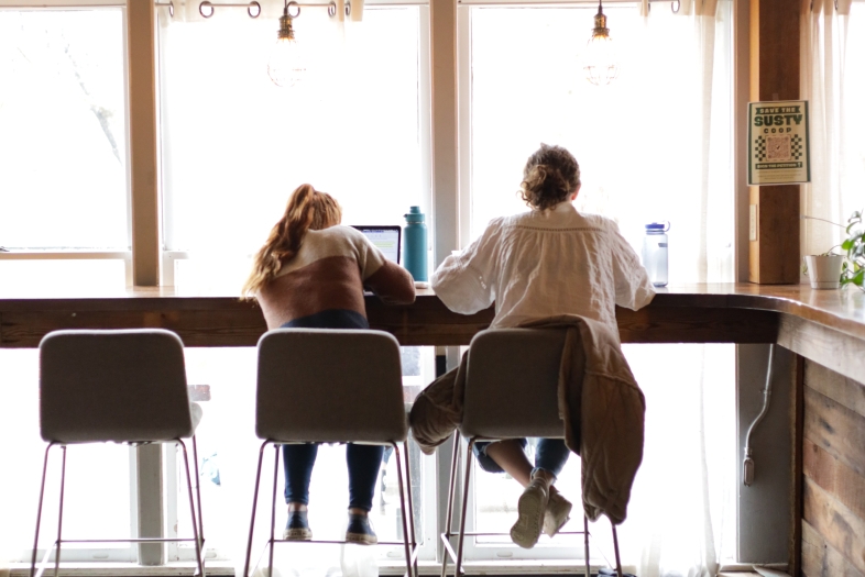 two students sit on high top stools while working on laptops by a window