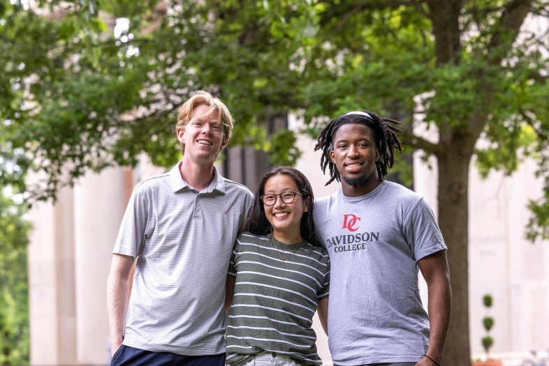 a group of three students standing together smiling outside