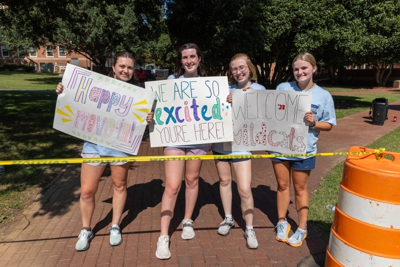 a group of four young women hold signs "welcome" to campus