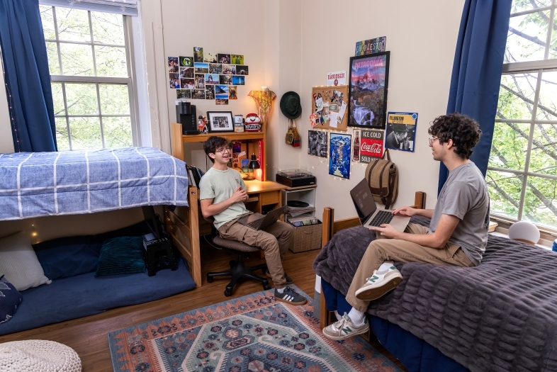 two young men sit in a dorm room talking and smiling with each other