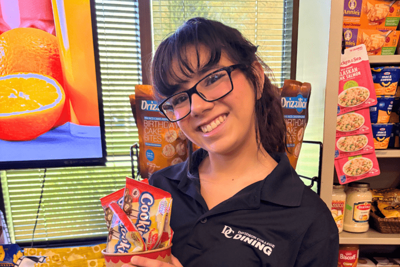 a young woman wearing a Davidson Dining shirt holds up snacks in a market