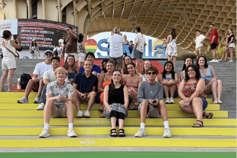 A group of approximately fifteen students sits on yellow and green steps branded with "Ayuntamiento de Sevilla" and "NO8DO" logos, positioned in front of the Setas de Sevilla wooden structure and a colorful "Sevilla" sign.