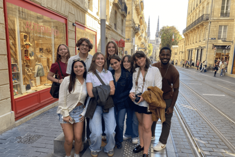 A diverse group of nine smiling students poses on a cobblestone street in Bordeaux next to an "Altermundi" storefront, with tram tracks and distant cathedral spires visible in the background.