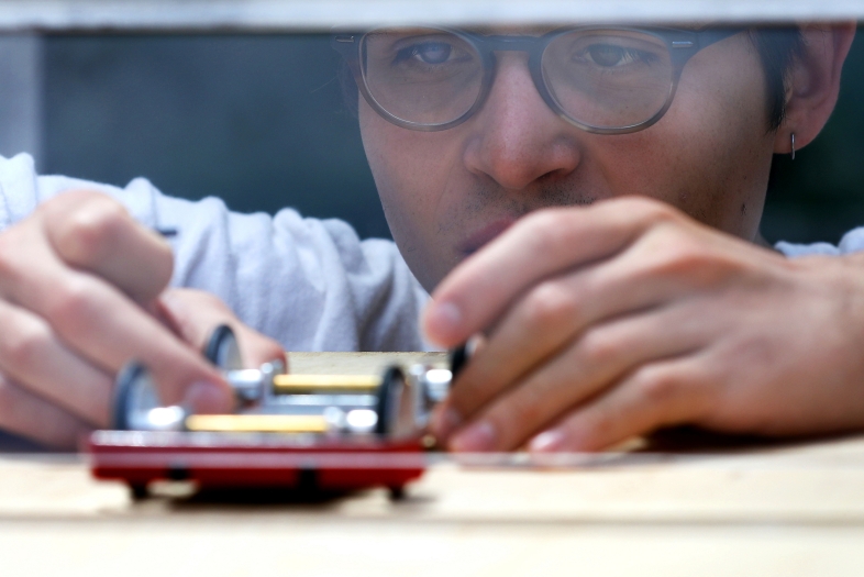 A close-up, eye-level shot shows a student wearing glasses leaning in with intense focus to adjust the wheels of a small model car on a wooden surface.