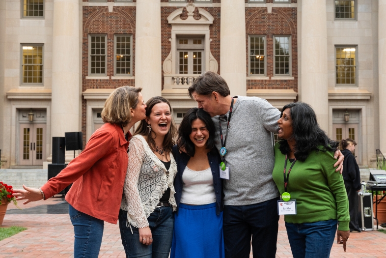 A group of five people laugh and embrace in a brick courtyard in front of a large, neoclassical building with white columns during Wildcat Weekend.