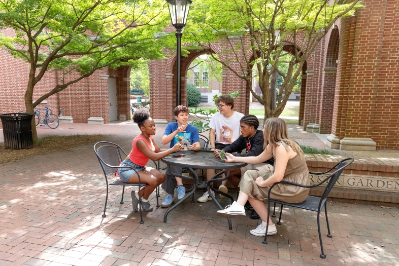 Five students of diverse backgrounds sit around a black metal mesh table in a brick courtyard, engaged in conversation under the shade of green trees and near a brick building with arched walkways.