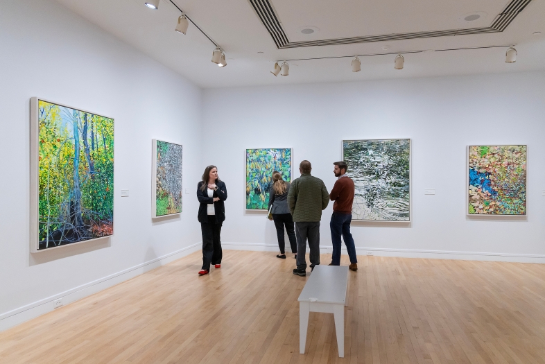 People view large, colorful Impressionist-style landscape paintings of forests and water in a brightly lit art gallery with white walls and light wood floors.