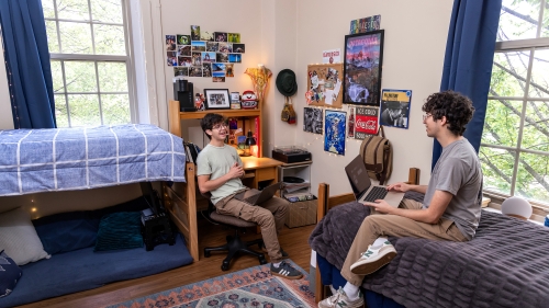 two young men sit in a dorm room talking and smiling with each other