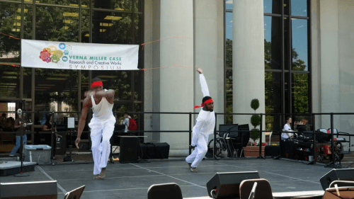 two young men dance on a stage outside of an academic building on a sunny day