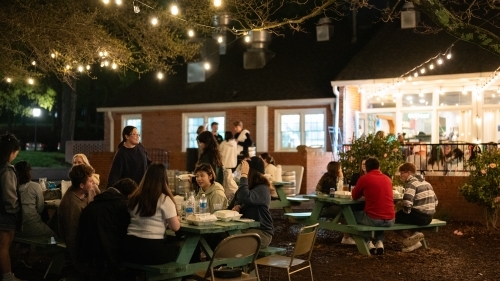 students sitting around picnic tables eating and smiling at night under string lights