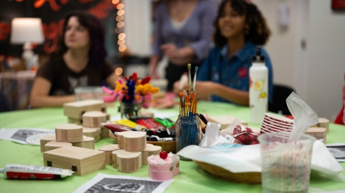 A green table is covered in art supplies, including unfinished wooden heart-shaped boxes, jars of paintbrushes, and acrylic paint tubes, while students gather in the blurred background of the studio.