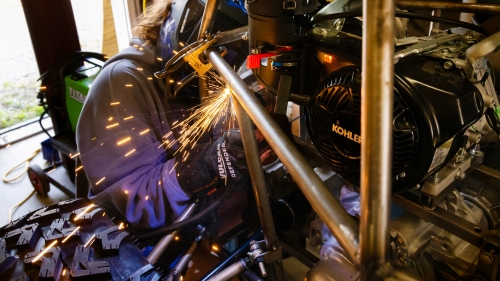 Close-up of a student wearing a welding helmet and safety gloves, working on a vehicle's metal frame near a Kohler engine as bright sparks fly out.