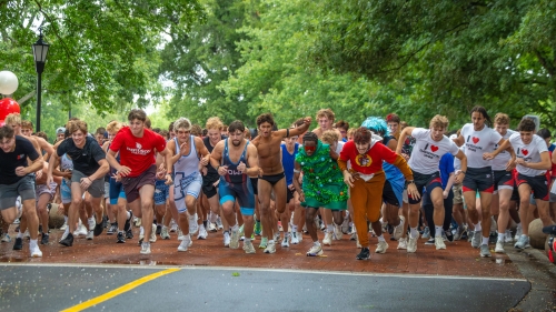 A large group of young men in athletic gear and costumes sprint down a brick paved street lined with green trees.