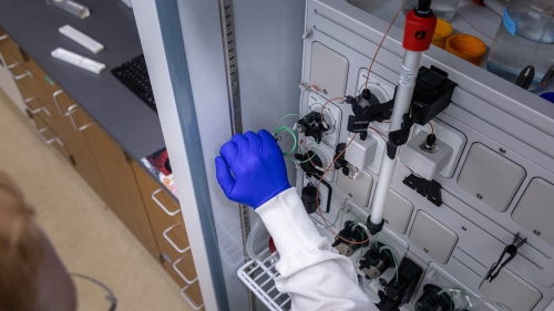 A researcher wearing a white lab coat and blue protective gloves adjusts the tubing on a laboratory machine inside a chromatography cabinet.