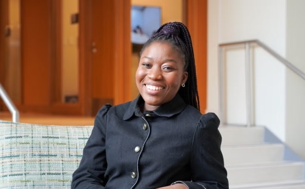 a young Black woman wearing a black jacket sits at a couch smiling