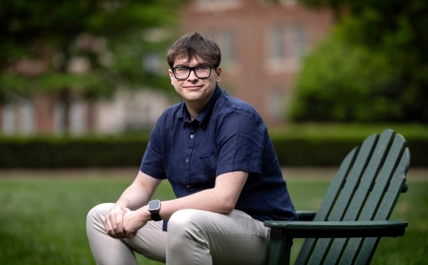 a young white man with a collared blue shirt sitting on a green adirondack chair on a college campus