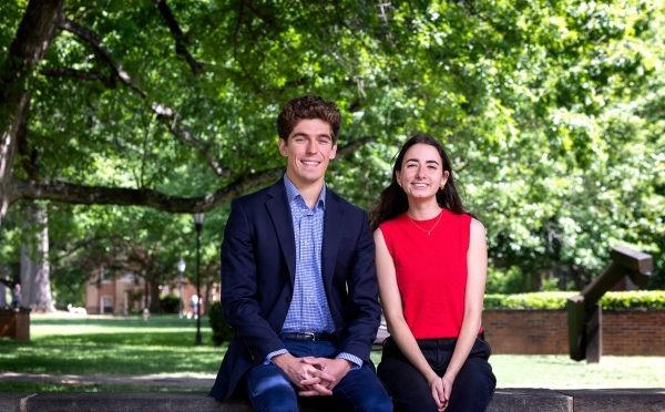 a young man and a young woman sit together on a brick wall in front of green trees