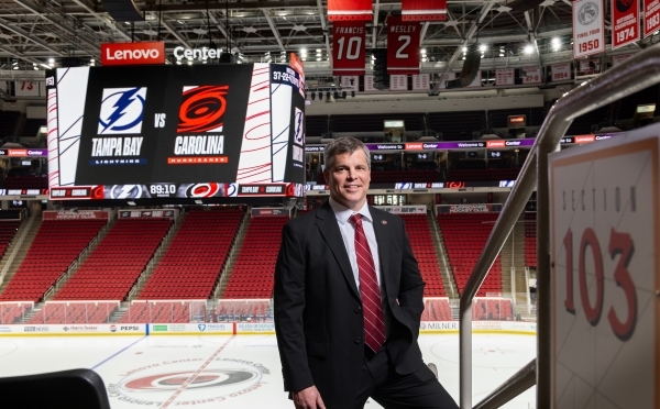 a middle aged white man wearing a black suit with a red tie in front of the Carolina Hurricanes hockey rink