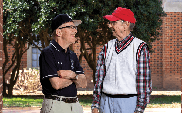two older men stand together smiling looking at each other in front of a stadium, wearing Davidson College gear
