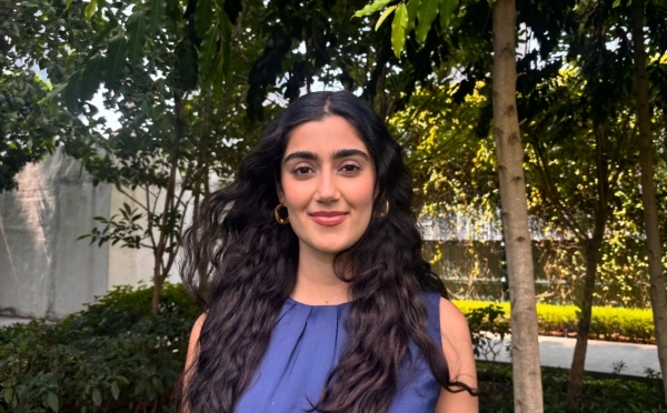 a young woman with long dark curly hair wearing a blue top standing outside in front of a set of green trees