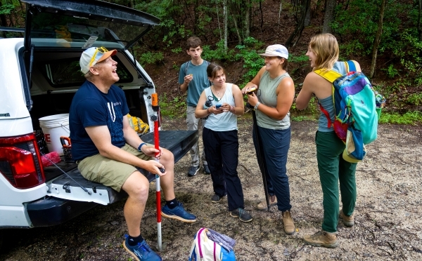 a group of students with their professor gathered around a truck in the woods wearing hiking gear