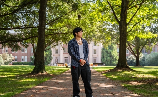 a young man stands on a college campus looking to his side in front of a large building surrounded by trees