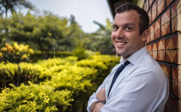 a man in a doctor's jacket smiles leaning against a brick wall