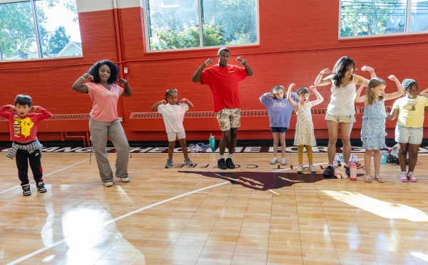 A group of diverse children and young adult leaders stand in a row on a gymnasium floor, smiling and flexing their muscles in a strong pose against a red and white wall.