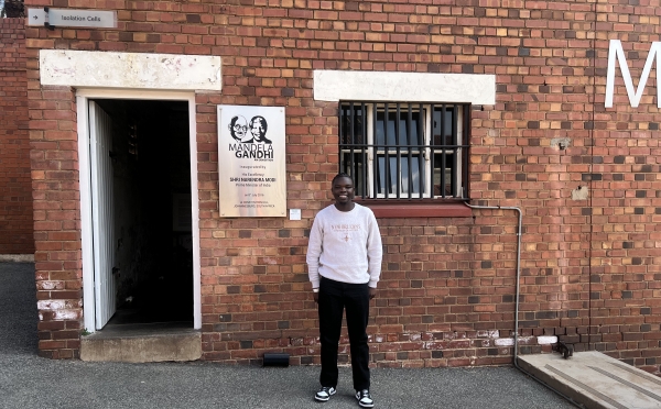 A man in a light grey sweatshirt and black pants stands in front of a red brick building at Constitution Hill in Johannesburg, next to an open doorway labeled "Isolation Cells" and a plaque commemorating the "Mandela Gandhi" exhibition.