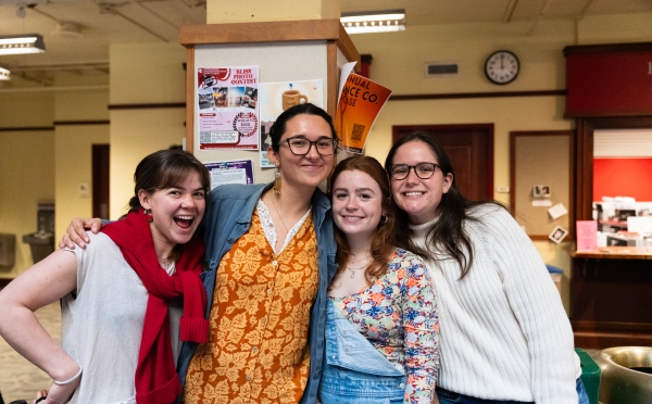 Four smiling young women pose closely together in a hallway decorated with community event posters.