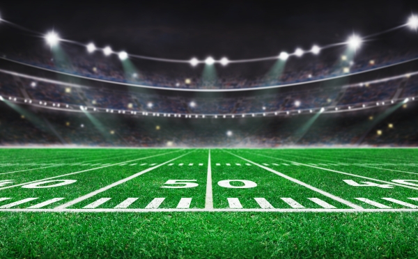 A wide-angle, low-altitude view of an American football field at night, focusing on the 50-yard line with a blurred stadium crowd and bright floodlights in the background.
