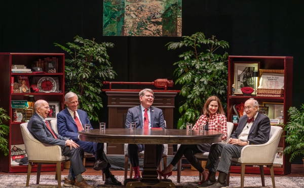 Five people sit around a large oval table on a stage set designed to look like a study, featuring bookshelves filled with memorabilia, potted plants, and an abstract painting hanging in the center.