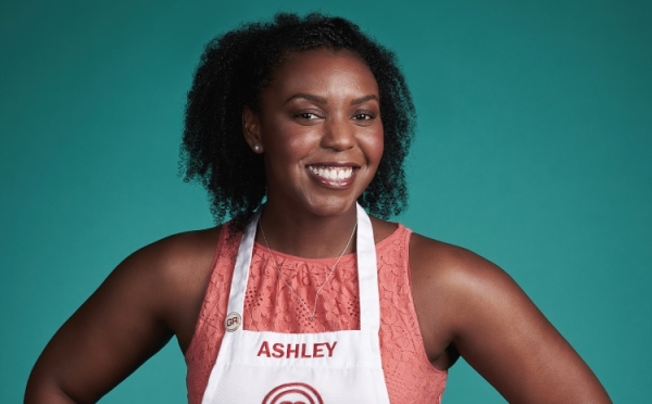 A smiling Black woman with curly hair wearing a white MasterChef apron with the name "ASHLEY" printed on it over a coral lace top, posed against a solid teal background.