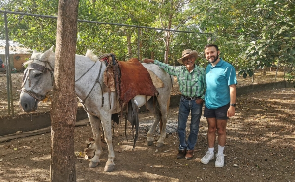 A man in a teal polo and an older man in a cowboy hat and flannel shirt stand together next to a saddled white horse in a fenced, sun-dappled yard.