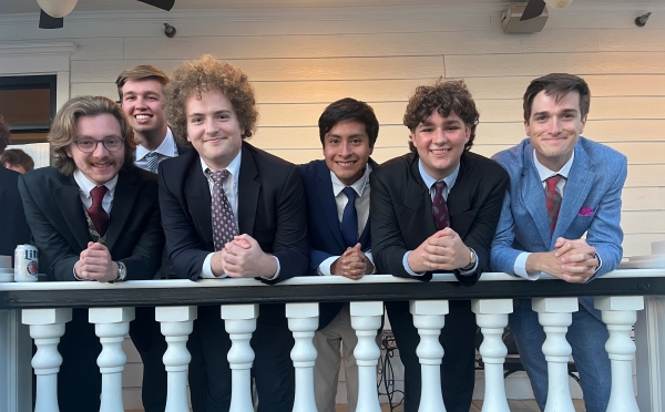 Six young men in formal suits and ties lean against a white balcony railing with classic balusters in front of a light-colored wooden house.
