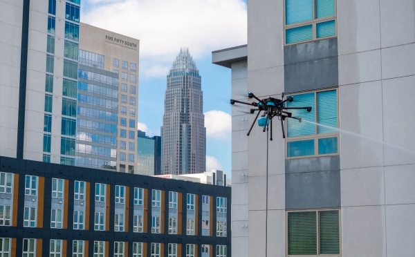 A professional-grade cleaning drone hovers beside a modern apartment building, spraying a stream of water onto the exterior windows against a city skyline featuring the Bank of America Corporate Center in Charlotte.