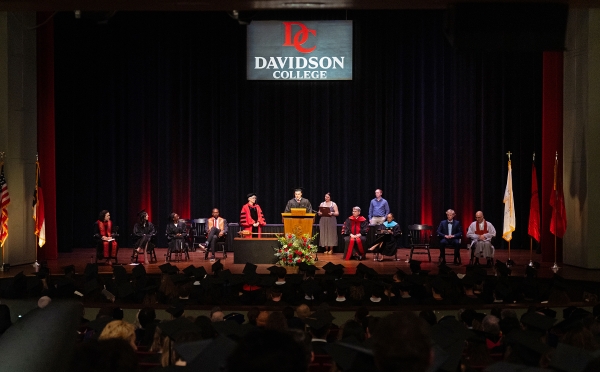 A wide view of a formal convocation ceremony on a stage with a "Davidson College" logo backdrop, featuring several faculty members in academic regalia seated around a student speaking at a podium