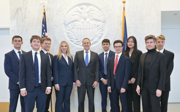 A group of ten professionally dressed students and a central official pose for a formal photo in front of a large, white architectural seal of the Federal Reserve Board of Governors.