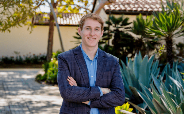A young white man wearing a blue blazer in a tropical setting, arms crossed