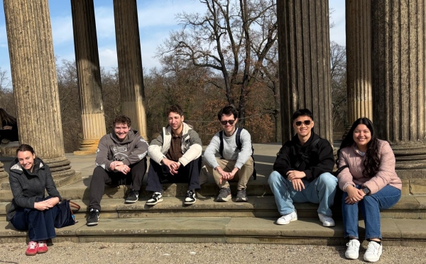 Six individuals sit smiling on the stone steps of a classical pavilion featuring large, fluted columns that frame a view of leafless trees under a clear blue sky.