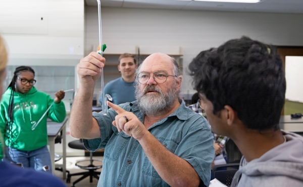 Professor Durwin Striplin, a bearded man wearing glasses, points to a piece of glass laboratory equipment while instructing a student in a chemistry lab setting.